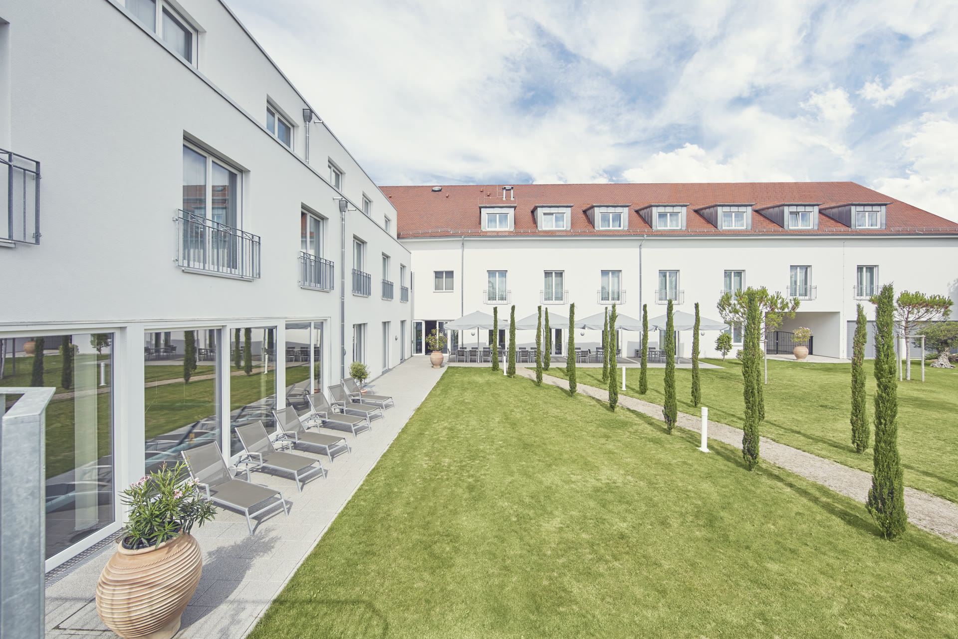 Begrünter Klinikgarten mit Zypressen, Wegen, Terrasse und Liegestühlen
