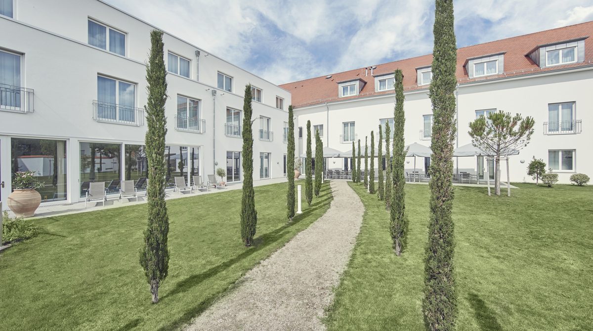 Innenhof der Schlossparkklinik Dirmstein mit Rasenfläche, Zypressen und einem Weg entlang der Klinikgebäude