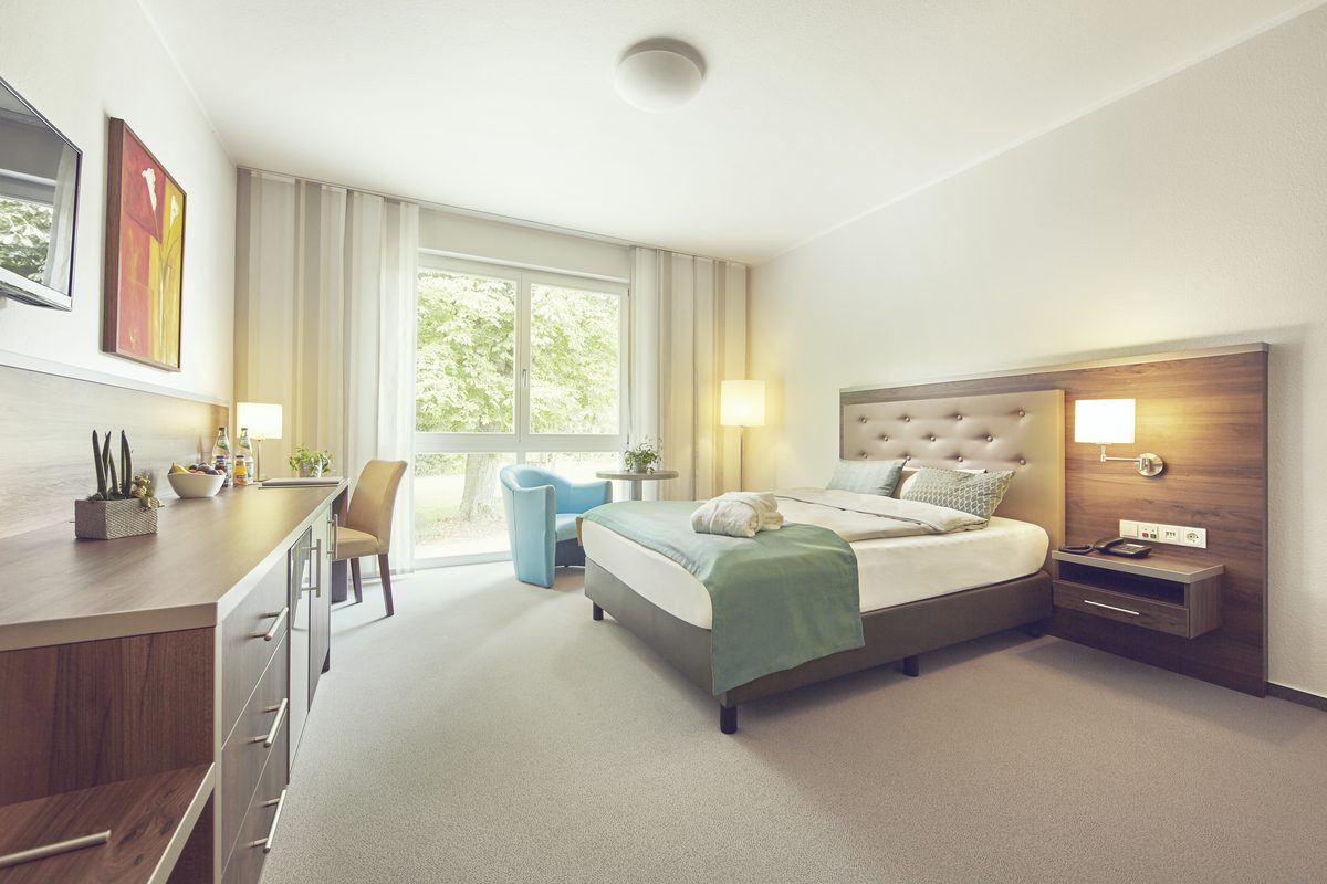 Patientenzimmer mit Boxspringbett, Schreibtisch, Sessel und Fenster zum Grünen